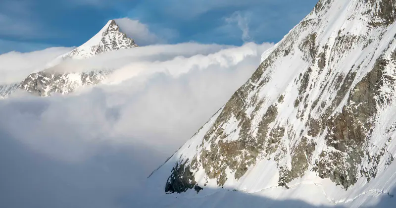 Een met sneeuw bedekte bergtop die boven de wolken uitsteekt, met op de voorgrond een steile, rotsachtige helling en op de achtergrond een blauwe lucht.